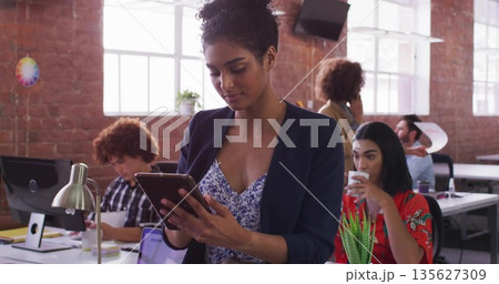 Standing South Asian woman in floral blouse and blazer using tablet in open office, with monitors 135627309