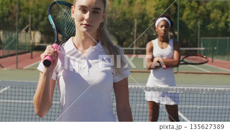 Standing tennis player wearing white gear shouldering racket on outdoor court, with net, fence 135627389