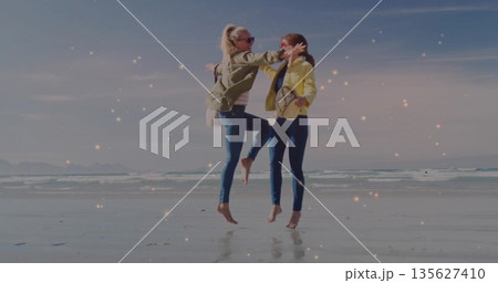 Leaping friends wearing olive and yellow jackets on beach near water's edge, with crossbody purses Leaping friends wearing olive and yellow jackets on beach near water's edge, with crossbody purses 135627410