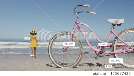 Woman wearing yellow sundress, straw hat standing facing waves on beach, with pastel pink bicycle Woman wearing yellow sundress, straw hat standing facing waves on beach, with pastel pink bicycle 135627426