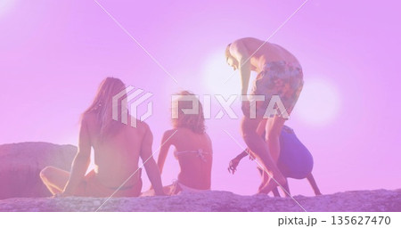 Sitting and climbing four friends in swimsuits on rocky outcrop at beach sunset, with ocean water 135627470