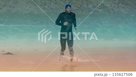 Athlete in black wetsuit running onto rocky lakeshore from shallow water, with swim cap and goggles Athlete in black wetsuit running onto rocky lakeshore from shallow water, with swim cap and goggles 135627506