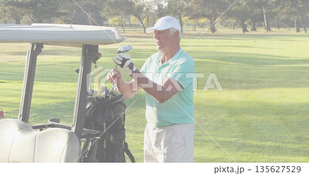 Senior male golfer wearing polo and cap selecting club from bag beside cart on fairway Senior male golfer wearing polo and cap selecting club from bag beside cart on fairway 135627529