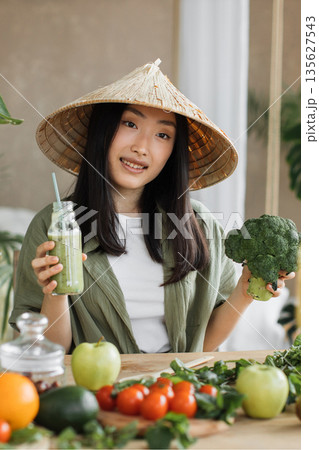 A young Asian woman wearing a conical hat holds a green smoothie and a head of broccoli, surrounded by fresh fruits and vegetables 135627543