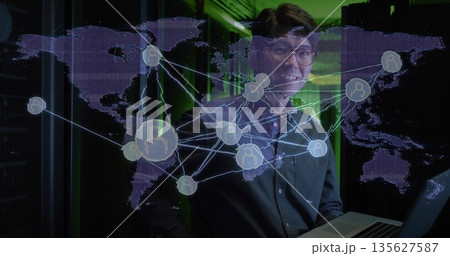 Standing IT professional in dark shirt holding laptop in green server room, with world map overlay Standing IT professional in dark shirt holding laptop in green server room, with world map overlay 135627587