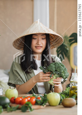 A young Asian woman wearing a conical hat prepares fresh broccoli and other vegetables for a healthy meal A young Asian woman wearing a conical hat prepares fresh broccoli and other vegetables for a healthy meal 135627765