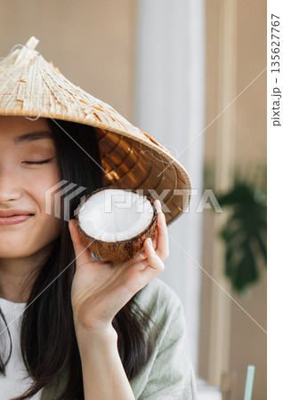 A young woman wearing a traditional conical hat holds a halved coconut near her face with a gentle smile 135627767