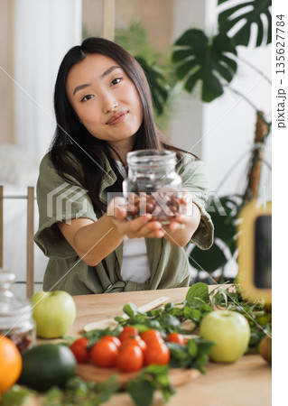 A young Asian woman smiles while presenting a jar filled with hazelnuts, surrounded by fresh fruits and vegetables on a wooden table, suggesting healthy eating and organic produce A young Asian woman smiles while presenting a jar filled with hazelnuts, surrounded by fresh fruits and vegetables on a wooden table, suggesting healthy eating and organic produce 135627784