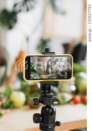 A young woman records a healthy food vlog on her smartphone, surrounded by fresh fruits and vegetables, showcasing a vibrant lifestyle 135627793