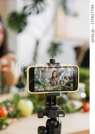 A young woman records a video about healthy eating and fresh ingredients using her smartphone on a tripod in a bright kitchen A young woman records a video about healthy eating and fresh ingredients using her smartphone on a tripod in a bright kitchen 135627794