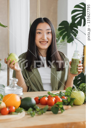 A young Asian woman smiles while holding a green apple in one hand and a bottle of green smoothie in the other, surrounded by fresh fruits and vegetables A young Asian woman smiles while holding a green apple in one hand and a bottle of green smoothie in the other, surrounded by fresh fruits and vegetables 135627795