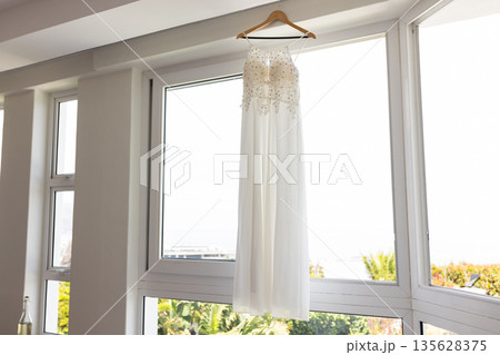 Elegant white wedding dress hanging by large window in bright room 135628375