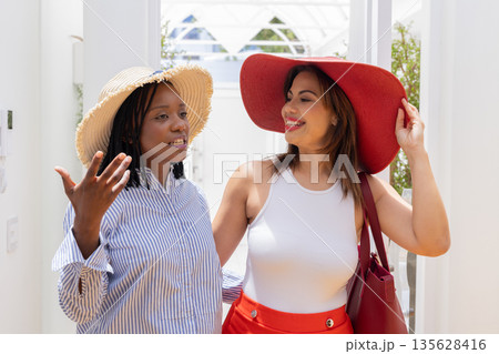 Two women wearing sun hats enjoying conversation at home, smiling and relaxed 135628416