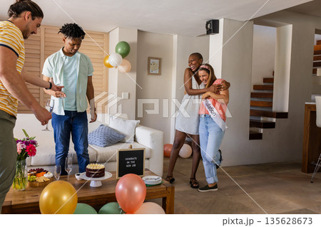 Friends celebrating engagement at home, hugging and smiling with cake and balloons 135628673