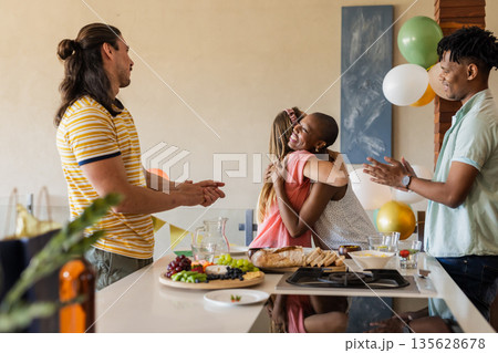 Friends celebrating at home, hugging and clapping around kitchen table with snacks 135628678