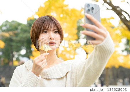 イチョウを背景にスマホで自撮りをするショートカットの女性 135628998