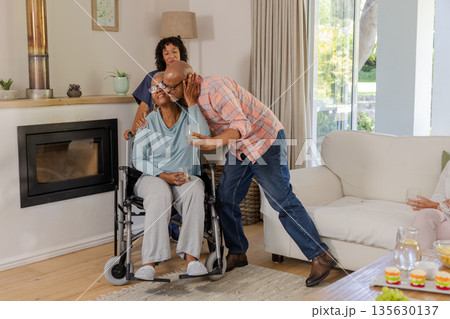 Elderly man kissing woman in wheelchair while friend smiles at home 135630137