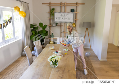 Senior woman decorating table for retirement party at home, arranging snacks Senior woman decorating table for retirement party at home, arranging snacks 135630141