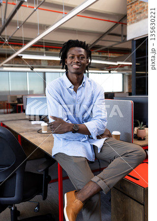 Smiling African American man sitting on desk in modern office, looking confident Smiling African American man sitting on desk in modern office, looking confident 135630221