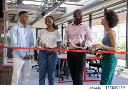 Colleagues celebrating office opening, smiling and chatting near red ribbon indoors 135630439