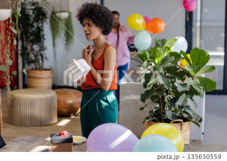 African American woman holding gift at office party with colorful balloons African American woman holding gift at office party with colorful balloons 135630559