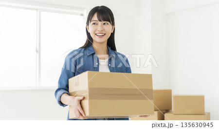 引っ越しの段ボール箱を抱えて笑顔で振り返るデニムシャツの日本人女性 135630945
