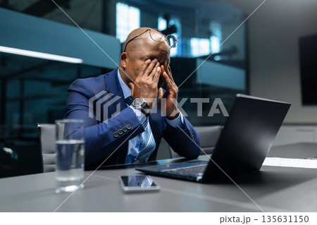 Exhausted mature black businessman rubbing eyes while sitting at desk in office Exhausted mature black businessman rubbing eyes while sitting at desk in office 135631150