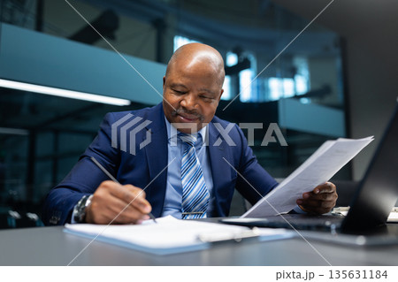 Confident African American businessman reviewing documents and signing paperwork at office desk Confident African American businessman reviewing documents and signing paperwork at office desk 135631184