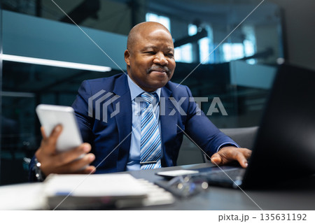 Smiling black mature businessman holding smartphone while working at desk in office 135631192
