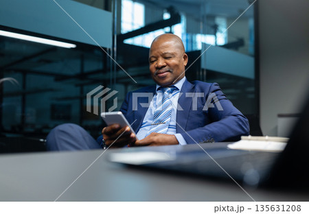 Confident black businessman using smartphone while sitting at workplace in office Confident black businessman using smartphone while sitting at workplace in office 135631208