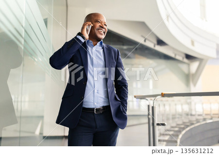 Smiling black mature businessman talking on smartphone while standing in office corridor 135631212