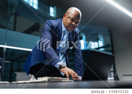 Focused black mature businessman leaning over conference table while working on laptop Focused black mature businessman leaning over conference table while working on laptop 135631232