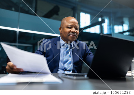 Confident black mature businessman reviewing paperwork while sitting at desk with laptop Confident black mature businessman reviewing paperwork while sitting at desk with laptop 135631258