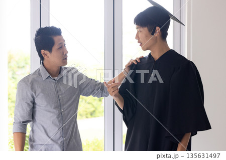 Celebrating academic achievement, father congratulating son in graduation gown at home Celebrating academic achievement, father congratulating son in graduation gown at home 135631497