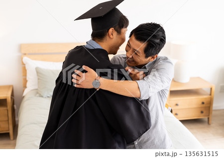 Graduation celebration, father hugging son in cap and gown at home Graduation celebration, father hugging son in cap and gown at home 135631515