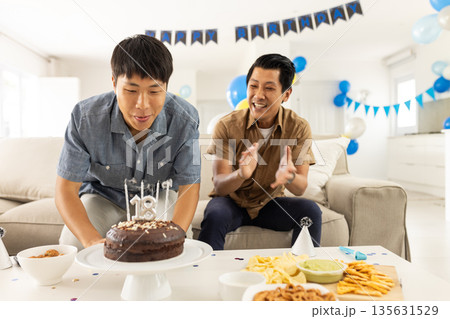 Blowing candles on birthday cake, Asian man with friend clapping joyfully Blowing candles on birthday cake, Asian man with friend clapping joyfully 135631529