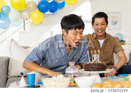 Celebrating birthday, young man blowing candles on cake with family at home 135631534