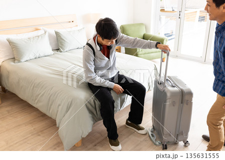 Young man sitting on bed with suitcase, preparing for travel at home 135631555