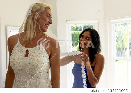 Bride in elegant gown smiling while friend adjusts veil in sunlit room 135631605