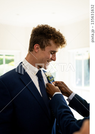 Groom smiling while friend adjusts boutonniere in bright wedding room 135631632