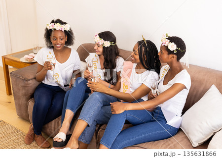 Toasting with champagne, diverse women wearing flower crowns celebrating friendship on sofa Toasting with champagne, diverse women wearing flower crowns celebrating friendship on sofa 135631866