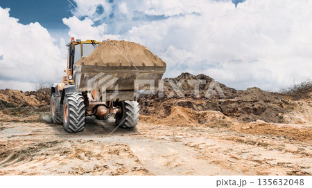 At a construction site, a loader moves soil to distribute it evenly, while nearby, under a partly cloudy sky, earthen embankments rise At a construction site, a loader moves soil to distribute it evenly, while nearby, under a partly cloudy sky, earthen embankments rise 135632048