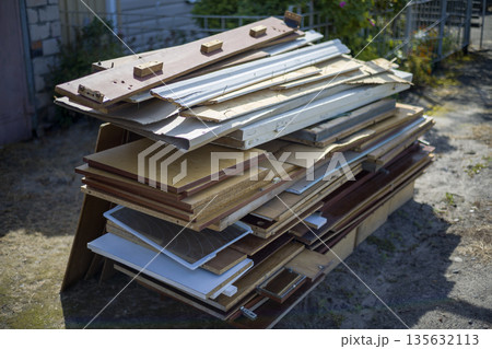 A stack of various wood sheets and boards rests against a fence in an outdoor space. The sun lights up the wooden materials on a clear day A stack of various wood sheets and boards rests against a fence in an outdoor space. The sun lights up the wooden materials on a clear day 135632113