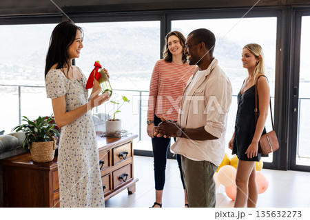Celebrating indoors, woman presenting champagne bottle with red ribbon to friends, smiling Celebrating indoors, woman presenting champagne bottle with red ribbon to friends, smiling 135632273