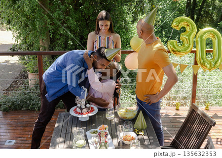 Celebrating birthday outdoors, friends hugging and smiling with cake and balloons 135632353