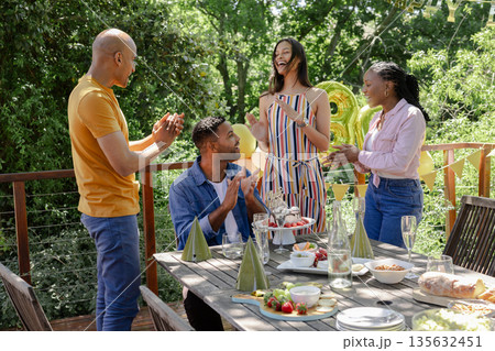 Friends celebrating birthday outdoors, clapping and laughing around decorated table 135632451