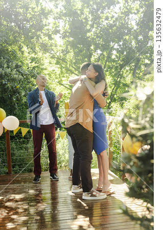 Friends celebrating outdoors, hugging and clapping on sunny wooden deck 135632479