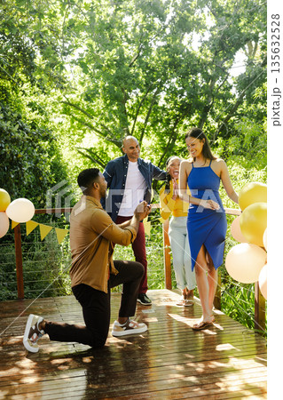 Man proposing to woman on sunny deck, friends celebrating with balloons Man proposing to woman on sunny deck, friends celebrating with balloons 135632528