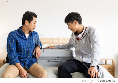 Two men sitting on bed packing suitcase, preparing for travel together Two men sitting on bed packing suitcase, preparing for travel together 135632613