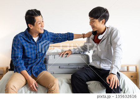 Father and son sitting on bed with suitcase, sharing joyful moment 135632616